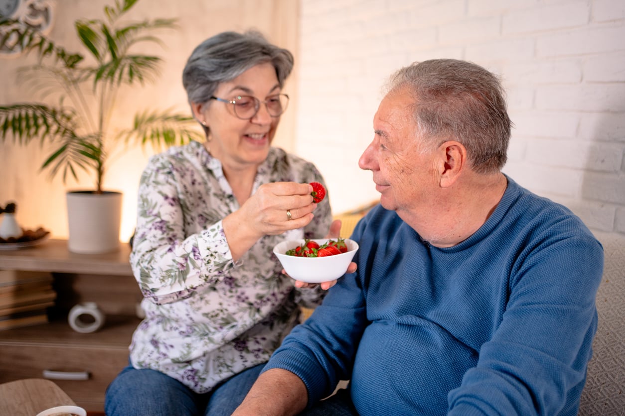 An older woman smiles as she offers a strawberry to an older man sitting beside her. She holds a bowl of strawberries, and they appear happy together in a cozy living room setting.