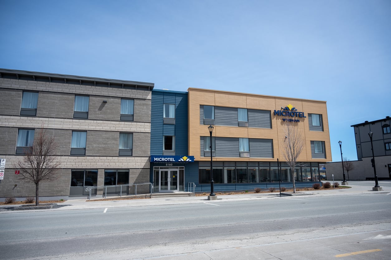A modern Microtel hotel building with three stories, large windows, and a mix of gray and tan exterior paneling. As one of the leading hotel chains, its contemporary design stands out along the empty street, where a few leafless trees line the sidewalk.