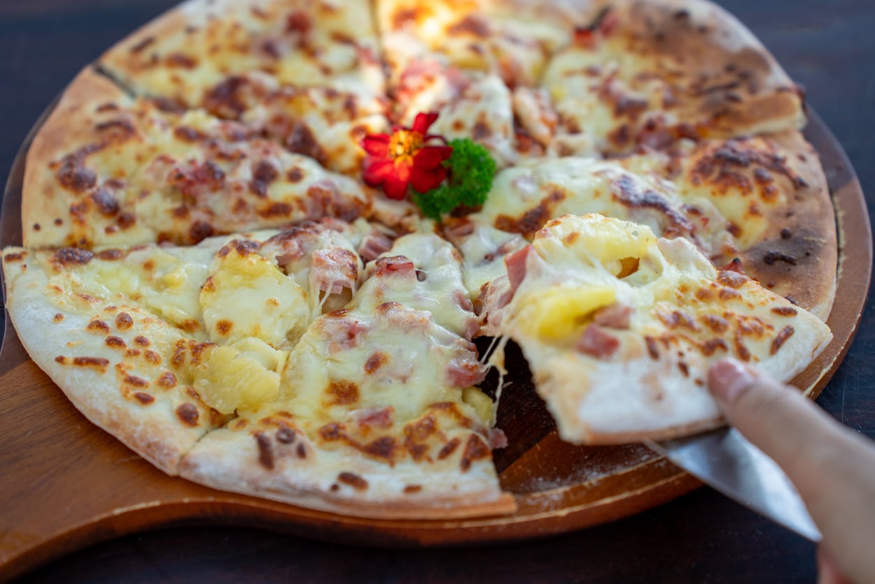 A hand lifts a slice of Hawaiian pizza with cheese, ham, and pineapple from a wooden board. A small red flower and parsley garnish the center of the pizza.