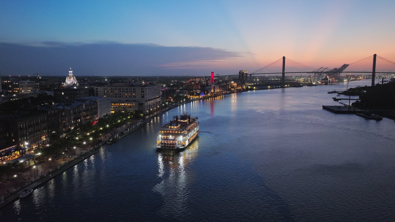 A riverboat cruises along a river at sunset, with city lights on the left, a bridge in the background, and a clear, colorful evening sky.