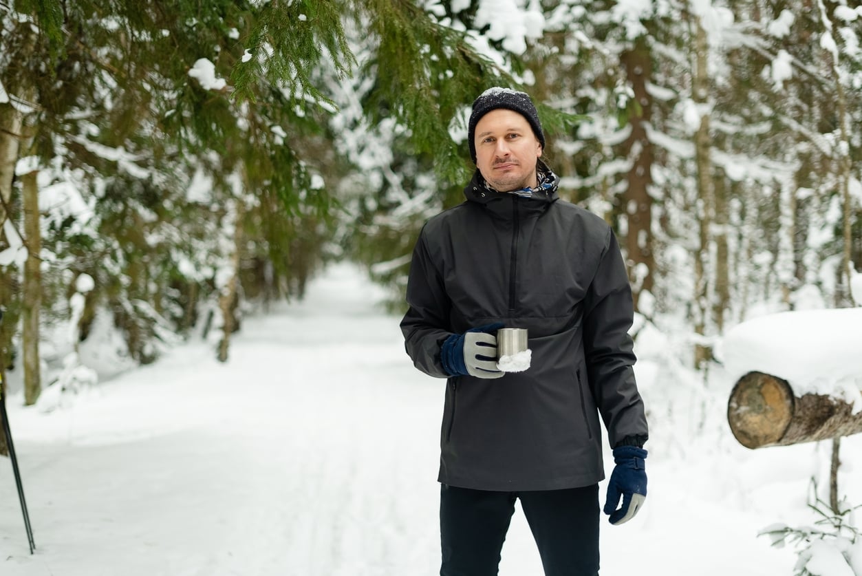 A person wearing a black jacket, gloves, and a beanie stands on a snowy forest path, holding a metal cup. Snow-covered trees line the trail.