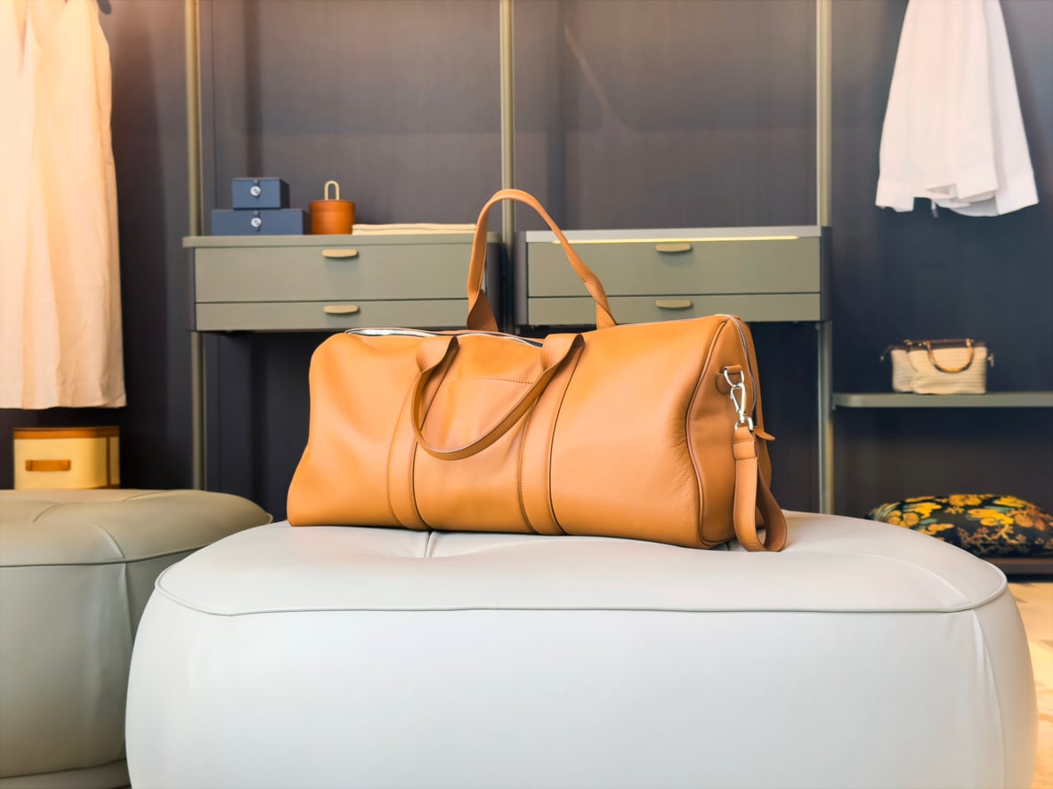 A tan leather duffel bag sits on a light ottoman in a modern walk-in closet with shelves, drawers, and hanging clothes in the background.