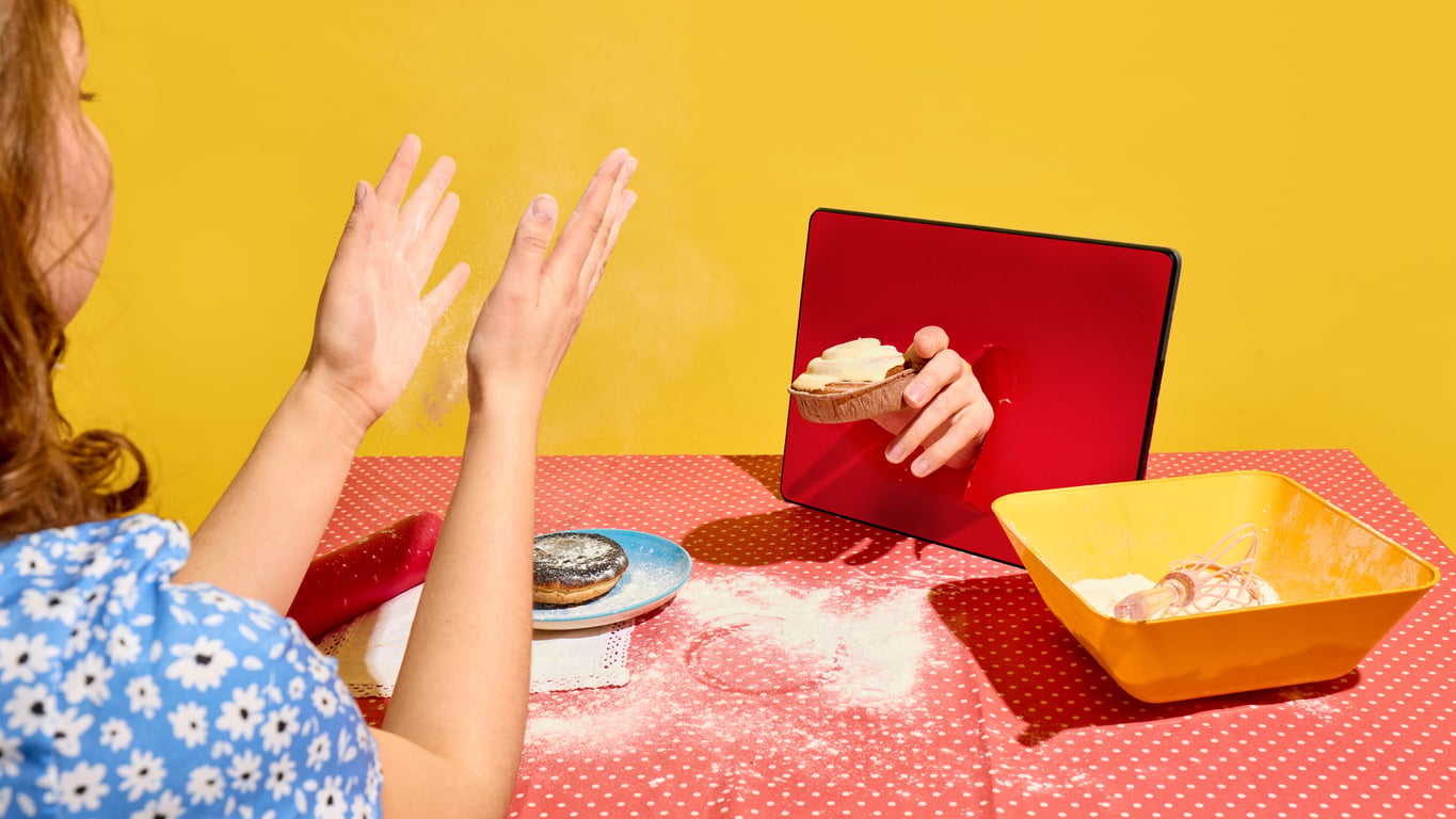 A person claps floury hands at a table while a hand reaches out from a tablet screen, offering a pastry. Baking tools and ingredients are scattered on the table.