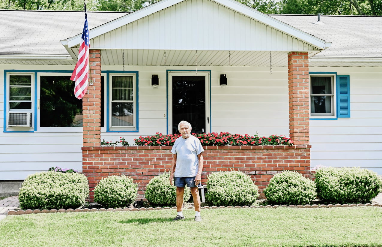 An older woman stands on the lawn in front of a white house with red brick columns, an American flag, and neatly trimmed bushes under a bright sunny sky.