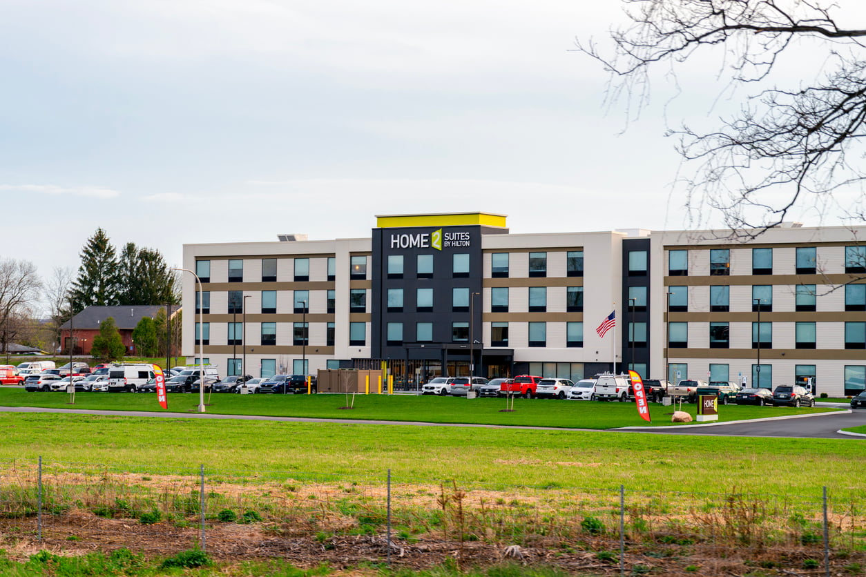 A modern three-story Home2 Suites by Hilton hotel, part of one of the leading hotel chains, with a parking lot full of cars, an American flag by the entrance, and green grass and trees in the foreground.