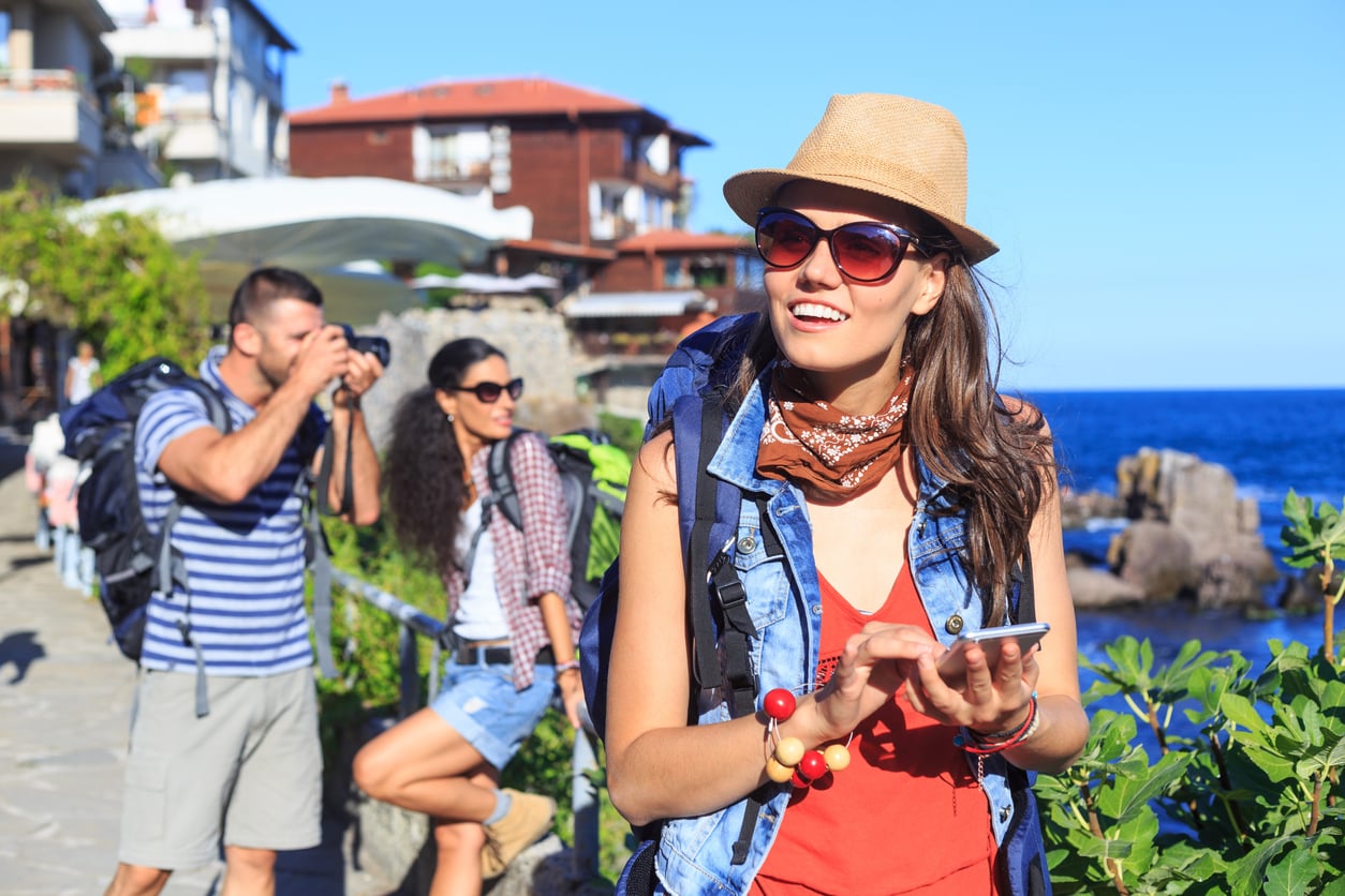 Three young adults with backpacks sightseeing by the sea; one woman smiles while using her phone, another woman poses, and a man takes photos. Sunny weather and scenic coastal buildings are in the background.