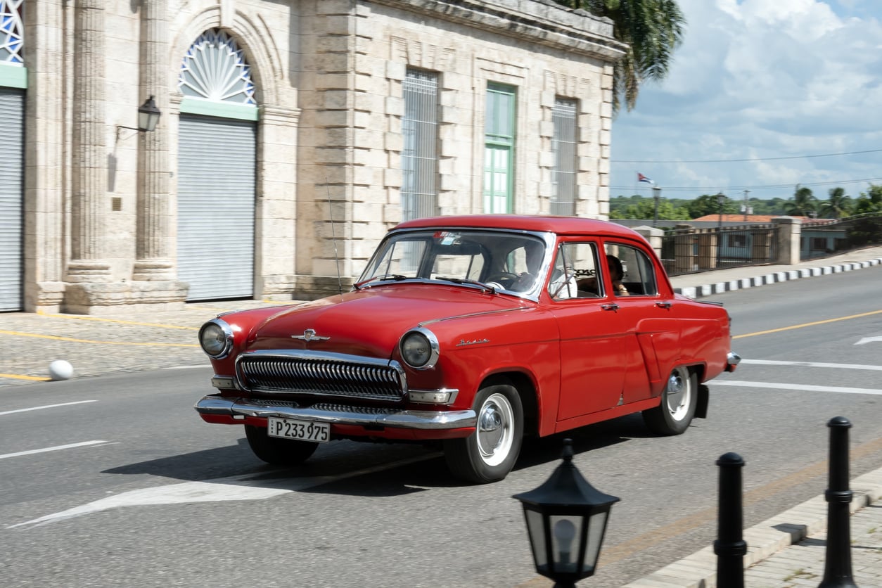 A vintage red car drives down a street lined with historic buildings and palm trees on a sunny day.