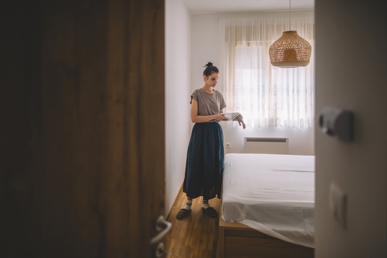 A woman stands in a bedroom near a neatly made bed, holding a book. The room has wooden floors, white walls, sheer curtains, and a woven pendant light hanging from the ceiling.