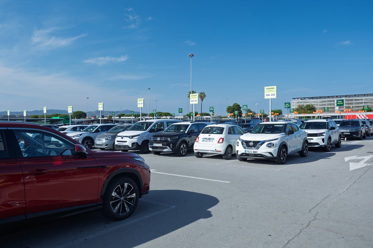 A large parking lot filled with various cars under a blue sky. Green and white signs are visible, and some greenery and buildings are in the background—a common scene where Vacation Mistakes like misplacing your car or forgetting where you parked can easily happen.