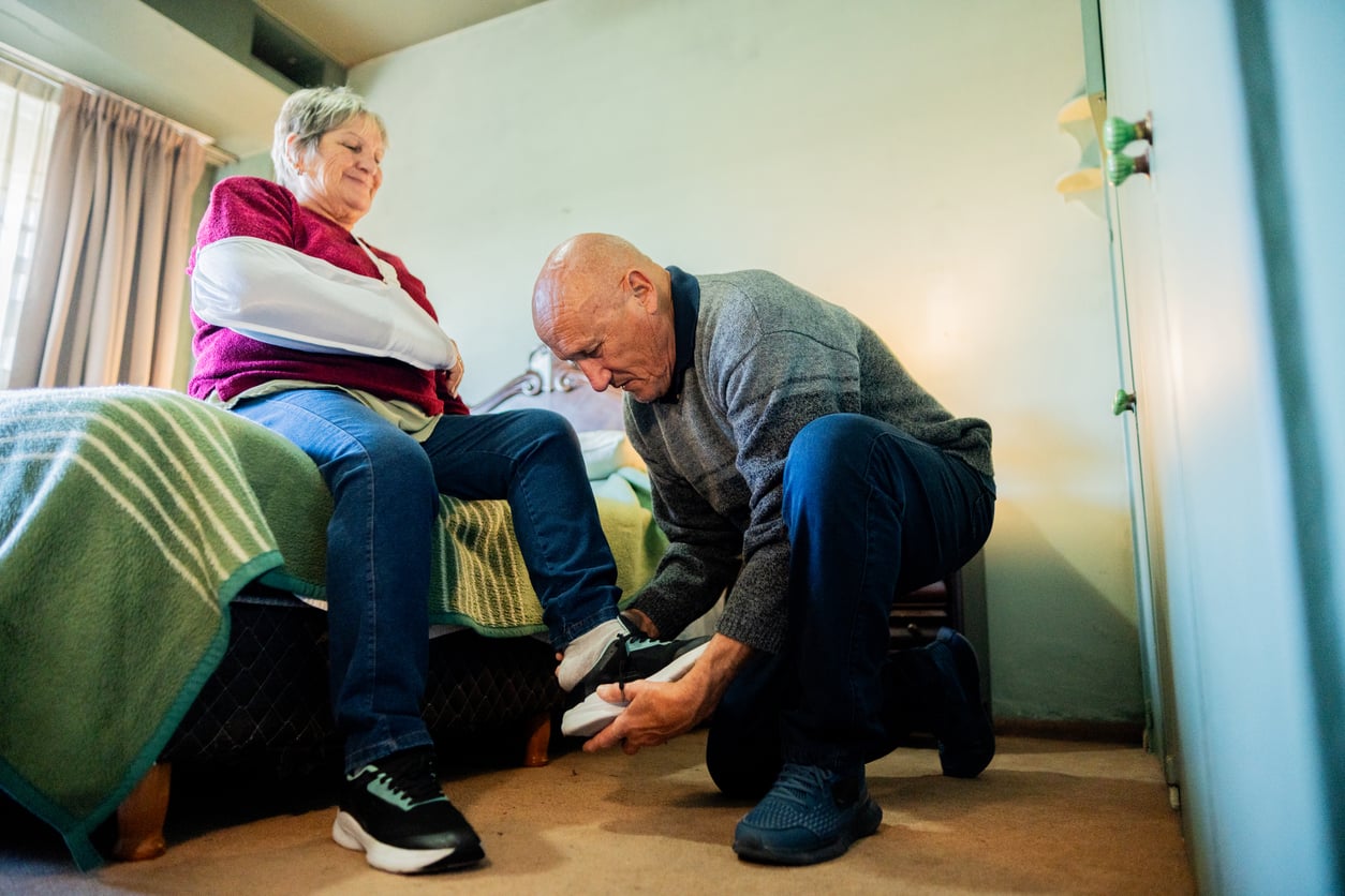 An older man kneels on the floor to help an older woman, who is sitting on a bed, put on her sneakers. They appear to be in a cozy, sunlit bedroom.