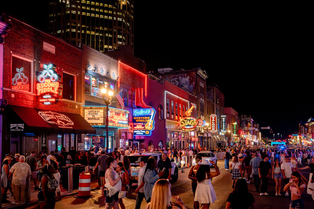 Busy city street at night with bright neon signs, crowded sidewalks, and people walking. Restaurants, bars, and music venues line the block, creating a lively, vibrant atmosphere.
