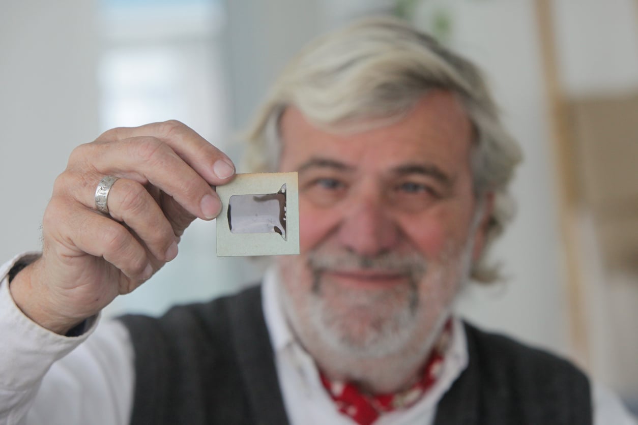 An older man with gray hair and a beard smiles while holding a photographic slide up to the camera, with the slide in clear focus and his face slightly blurred in the background.