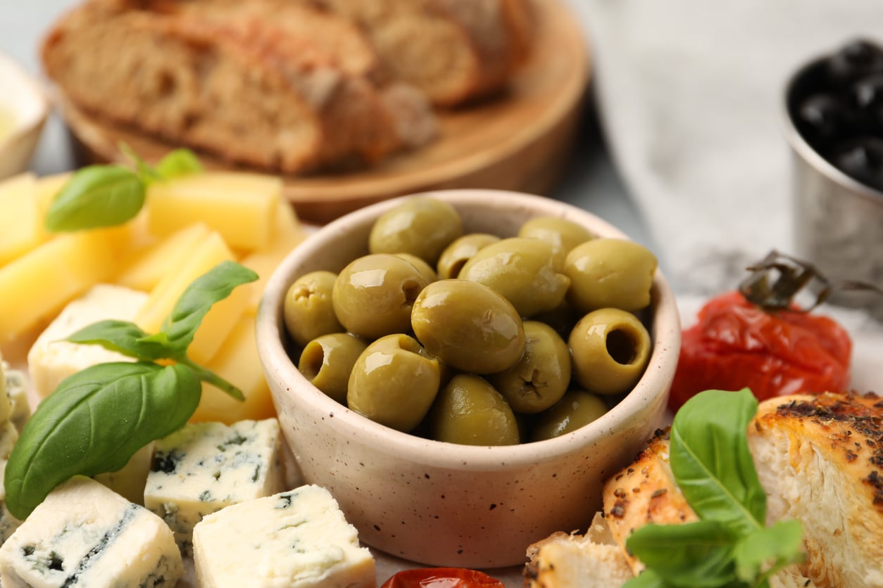 A bowl of green olives, one of those foods people learn to appreciate later in life, is surrounded by cheese cubes, blue cheese, fresh basil, grilled chicken, roasted tomato, black olives, and sliced bread on a wooden board.