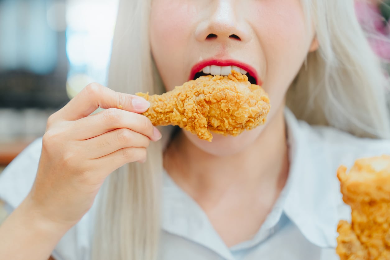 A person with long light hair and red lipstick is about to bite into a crispy fried chicken drumstick, holding it close to their mouth with one hand.