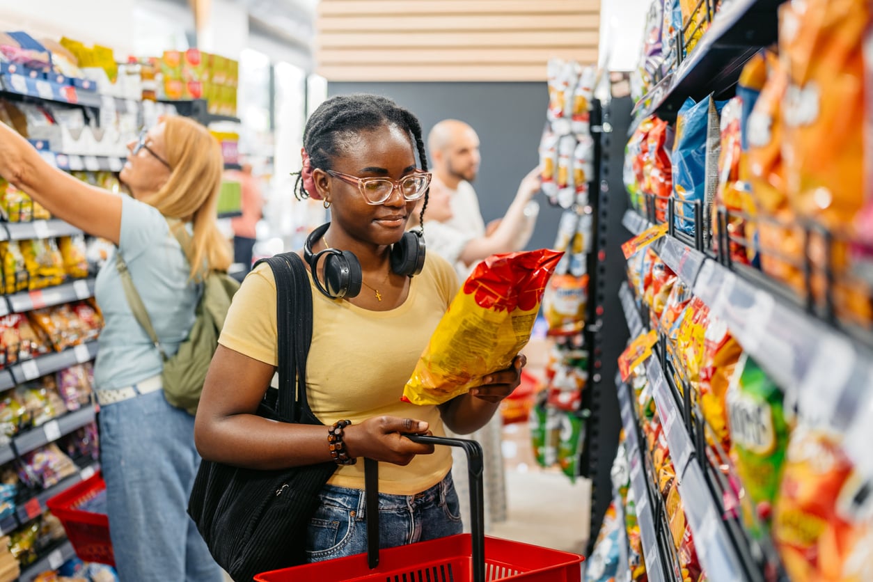 A person holding a shopping basket and reading a bag of chips in a grocery store aisle, with other people shopping in the background.