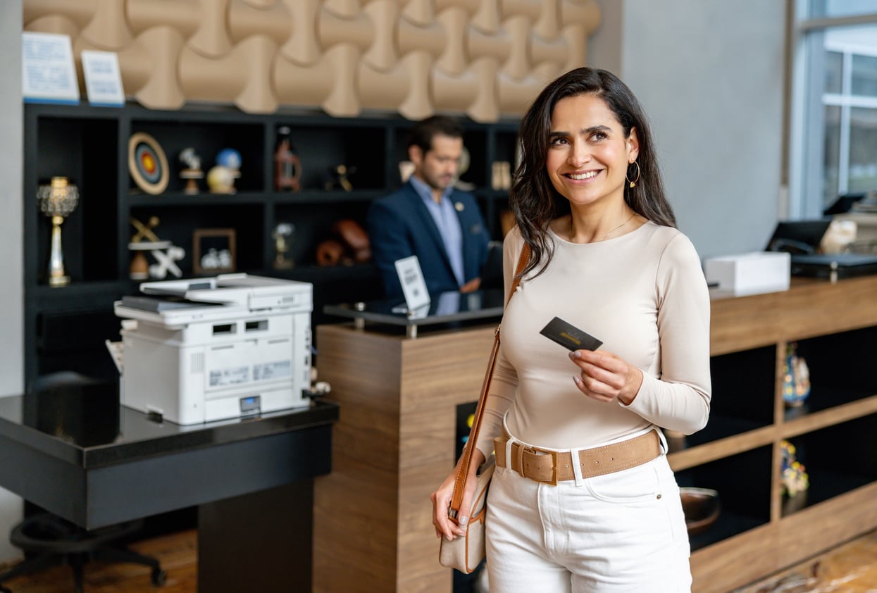 A woman holding a card and smiling stands in front of a hotel reception desk, while a receptionist works in the background.