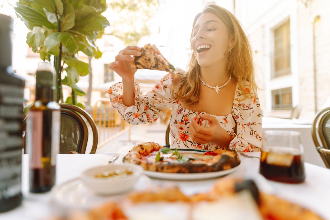 A woman in a floral dress smiles as she holds up a slice of pizza at an outdoor restaurant table, with pizza, a drink, and condiments in front of her. The setting is bright and sunny.