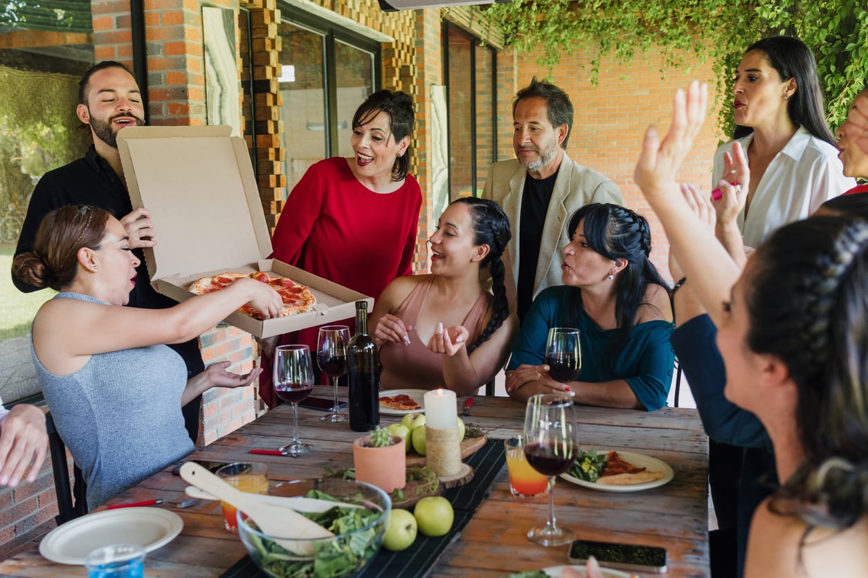 A group of people gathered around a wooden table outdoors, smiling and talking as one person opens a pizza box. Food, drinks, and fruit are on the table. Some people are reaching for pizza.