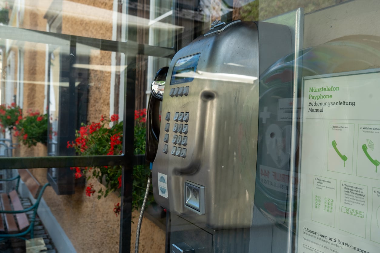 A silver payphone is mounted inside a glass booth, with red flowers visible outside. A sign with phone instructions in German and a diagram of a phone handset is posted on the right wall of the booth.