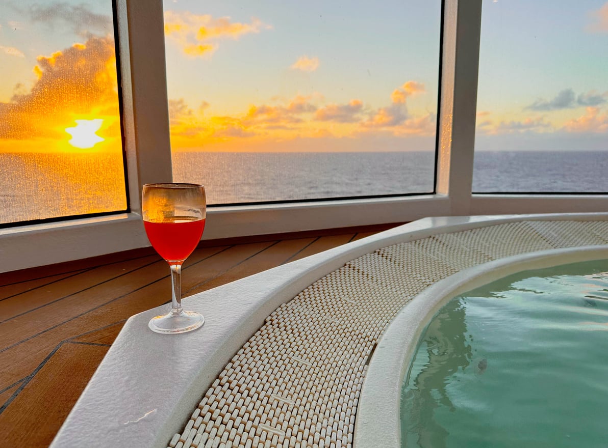 A glass of red drink sits on the edge of a hot tub with an ocean view at sunset, seen through large windows.