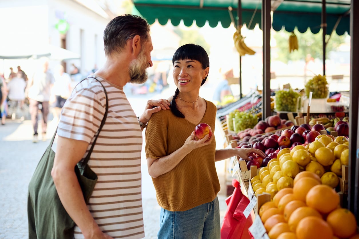 A woman holding an apple smiles and talks to a man at an outdoor fruit market, surrounded by apples, peaches, and oranges on display—capturing the essence of authentic vacation experiences.