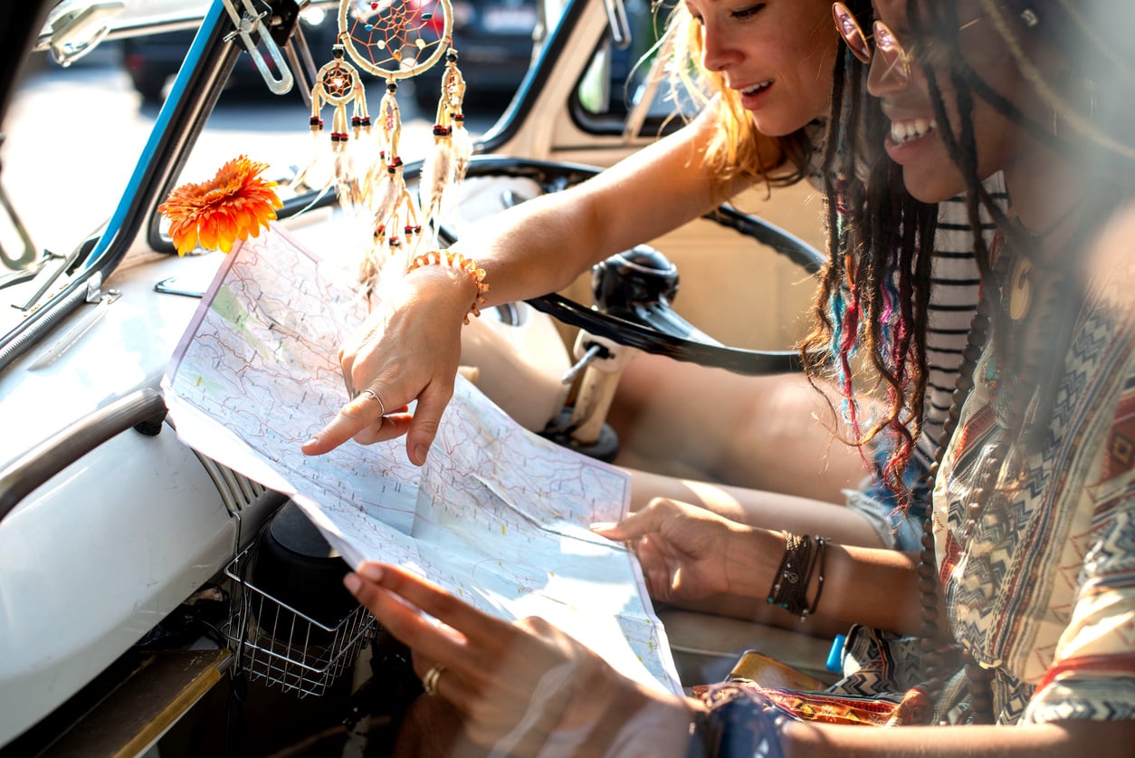 Two women sitting in a van look at a map together. Dreamcatchers and a flower hang from the rearview mirror, and sunlight streams inside. The mood is cheerful and adventurous.