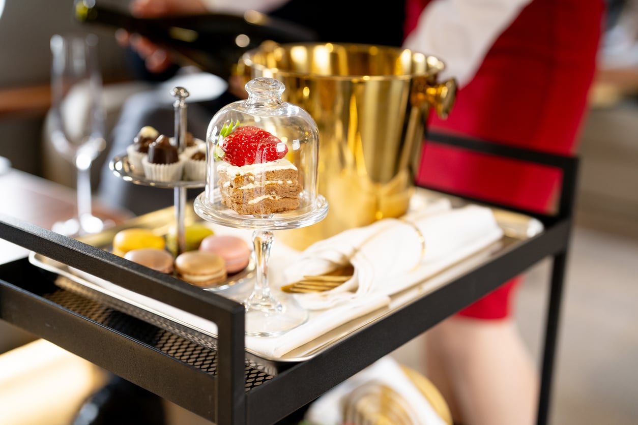 A dessert cart with assorted sweets, macarons, a glass dome covering a cake, a gold ice bucket, napkins, and a hand holding a bottle. The cart and items appear elegant and sophisticated.