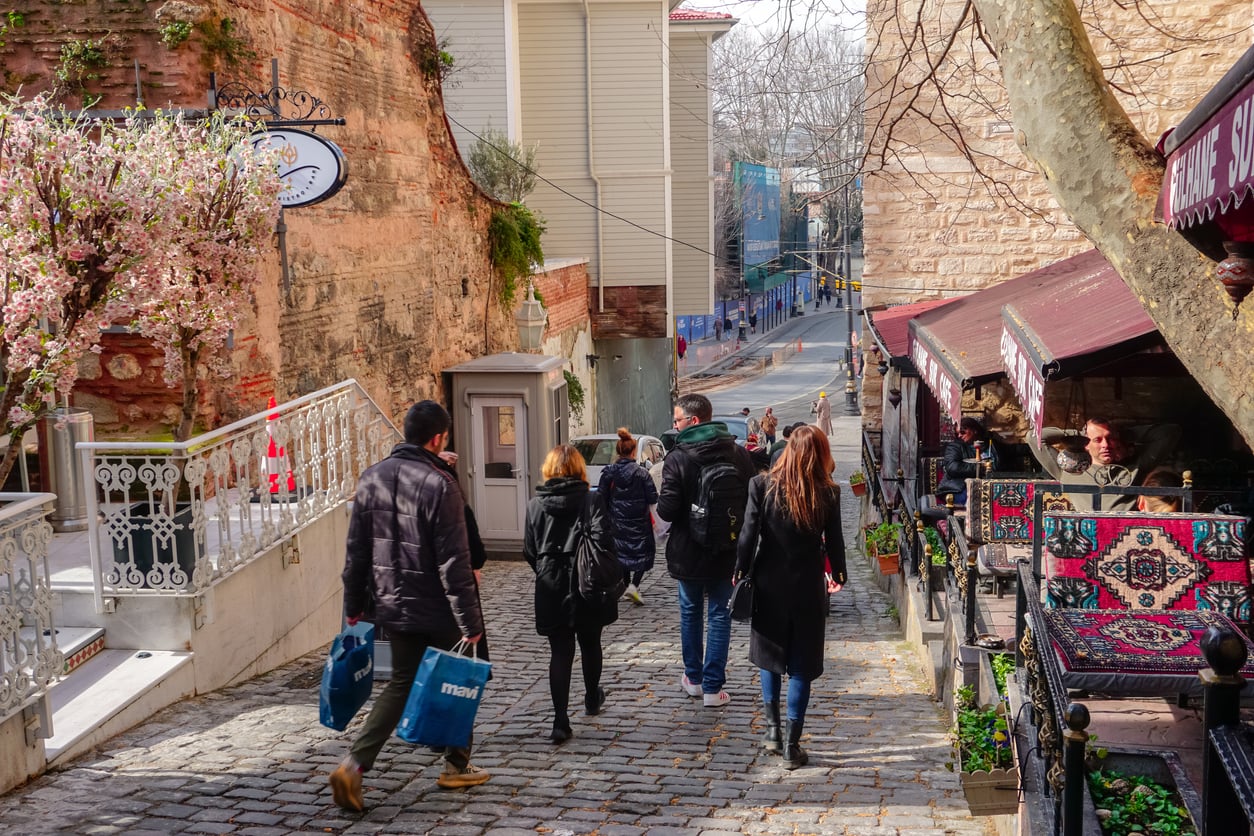 Five people walk down a cobblestone street lined with shops and blooming trees, careful not to let common vacation mistakes ruin their day. Outdoor seating and colorful rugs appear on the right, while the sun casts shadows over the vibrant, lively scene.