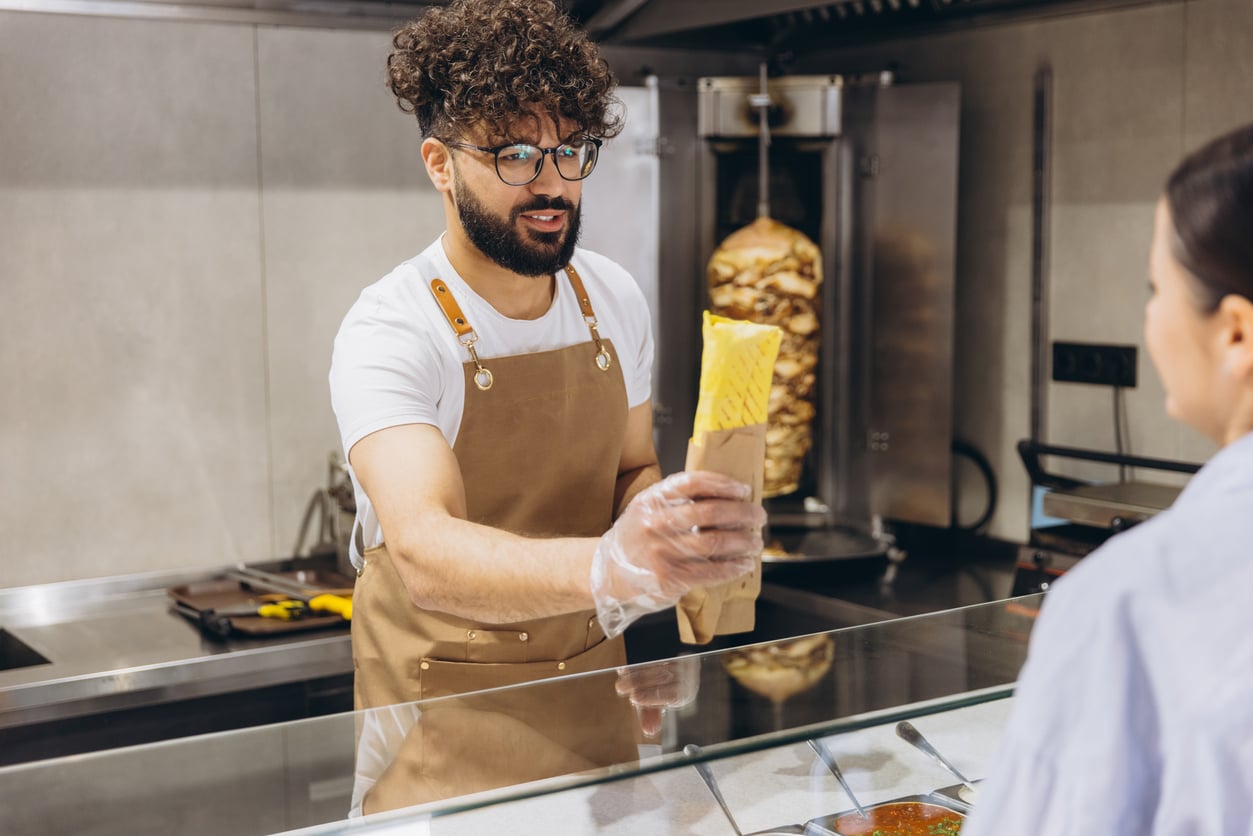 A man wearing an apron and gloves hands a wrapped sandwich to a customer at a food counter, with a vertical rotisserie visible in the background.