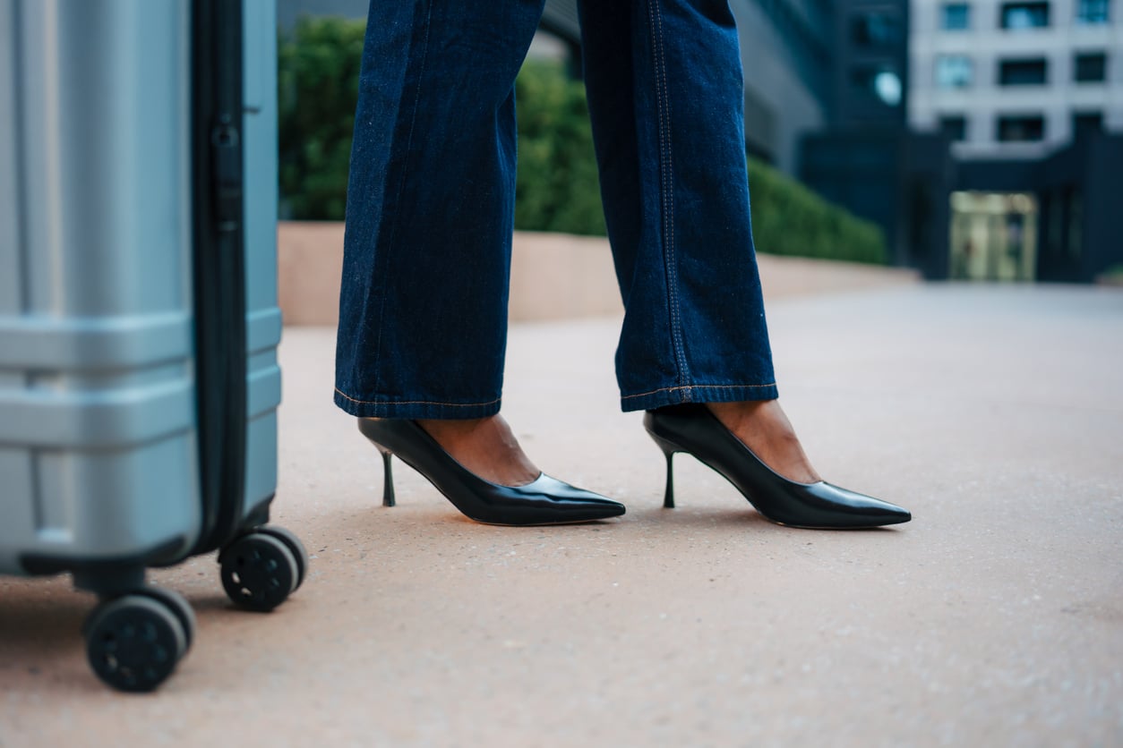 A person wearing black heels and blue jeans stands next to a silver rolling suitcase on a city sidewalk, with buildings and greenery in the background.