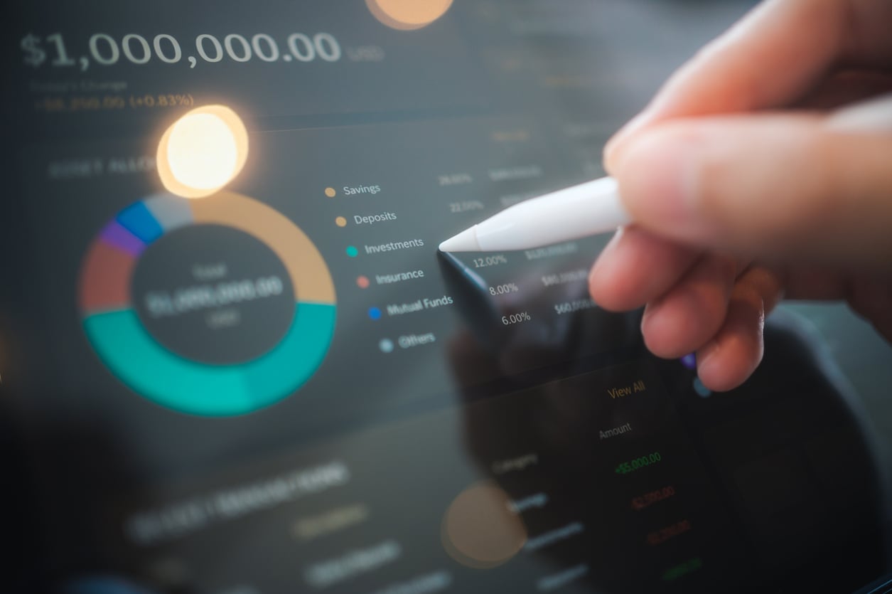A hand holding a stylus points at a tablet screen displaying financial graphs, pie charts, and investment data.