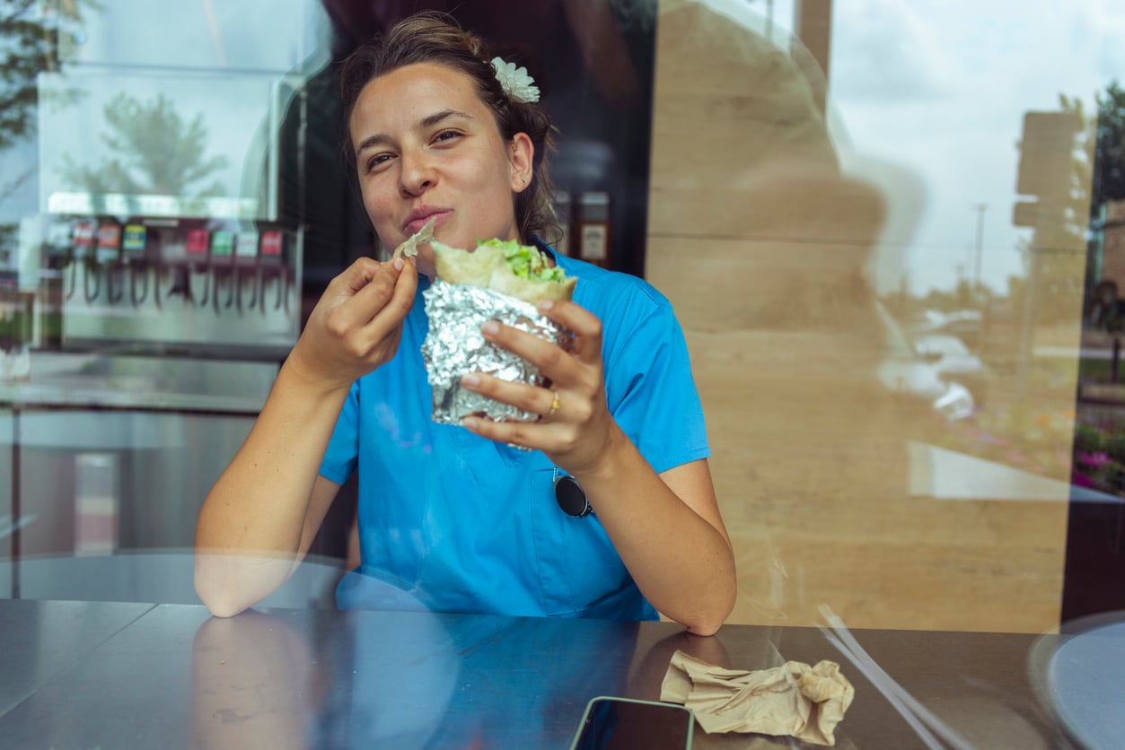 A woman in blue scrubs smiles and eats a large burrito wrapped in foil while sitting at a table by a window, with napkins and a phone nearby.