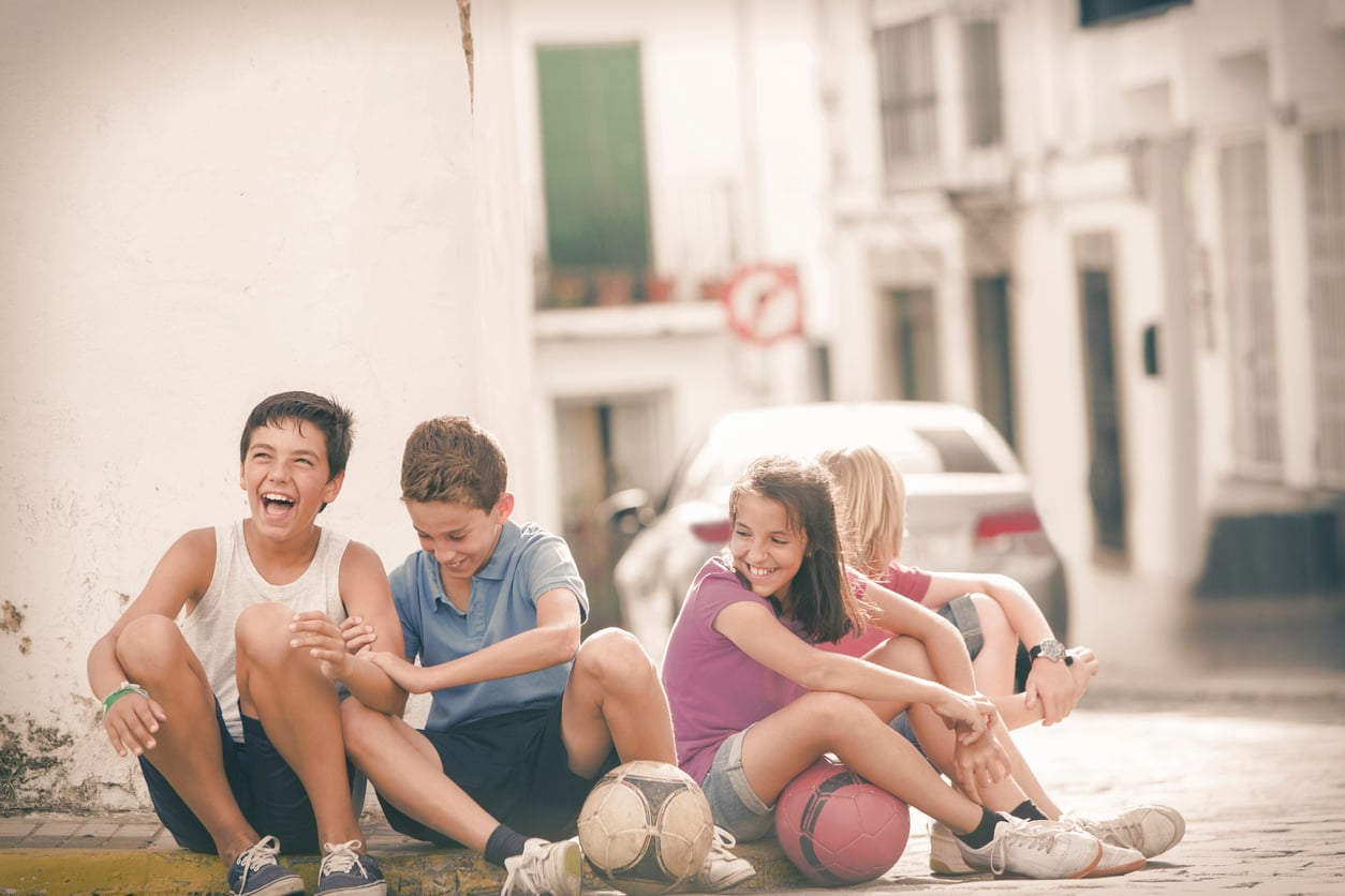 Four children sit on a sunny sidewalk, laughing and relaxing near two soccer balls. The background shows parked cars and white buildings, creating a playful, carefree atmosphere.