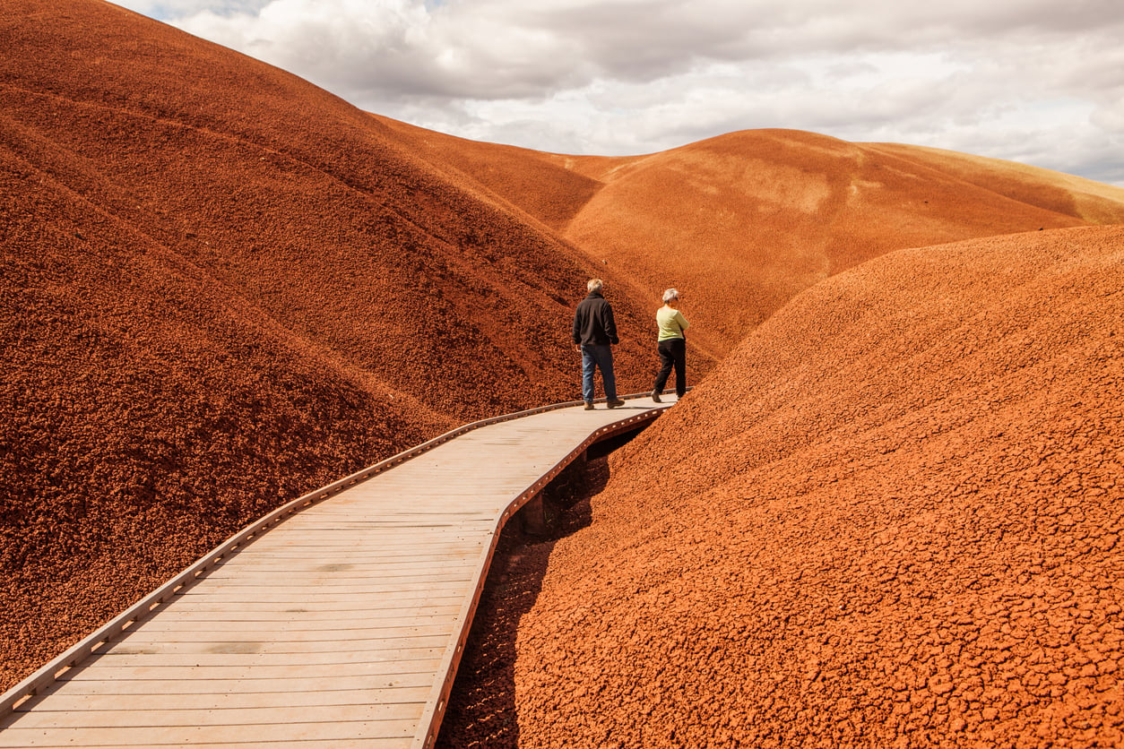 Two people walk along a wooden boardwalk winding through orange-red, barren hills under a partly cloudy sky.