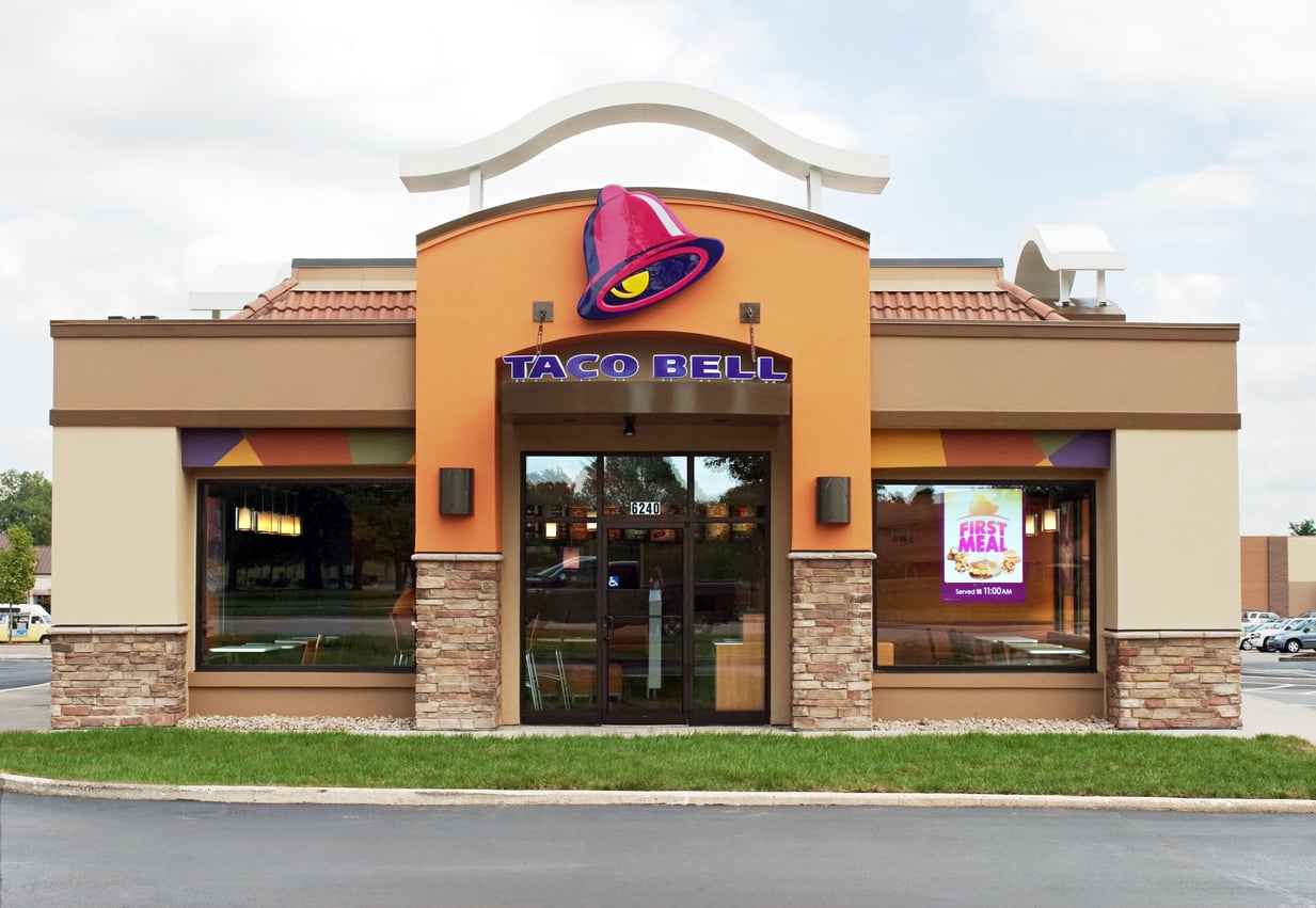 A Taco Bell restaurant with its signature bell logo above the entrance, large front windows, and a parking lot in front. The building has orange and brown tones with stone accents at the base.
