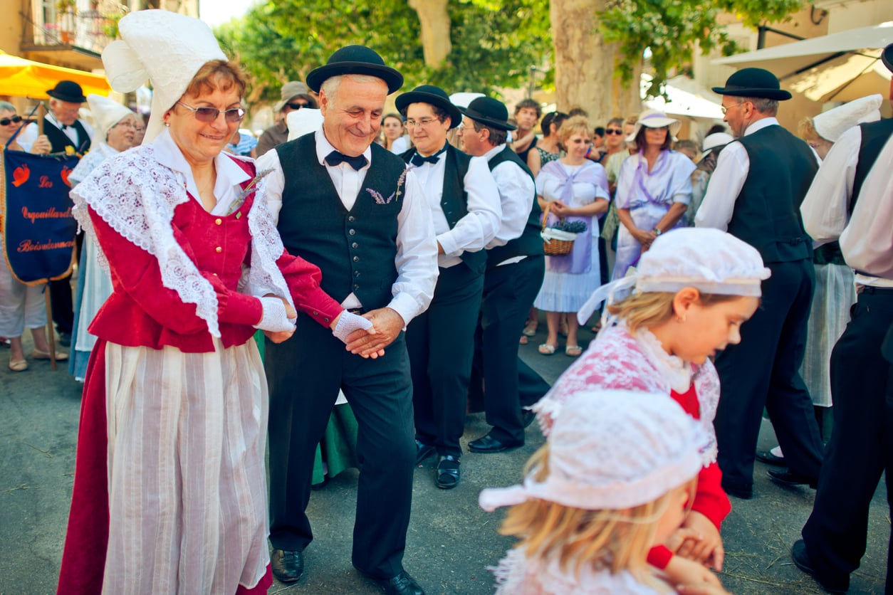 People in traditional European costumes dance outdoors in a festive parade, creating memorable vacation experiences for all who attend. Adults in lace and hats hold hands and smile, while children in matching attire walk in the foreground. A crowd watches in the background, enjoying the lively celebration.