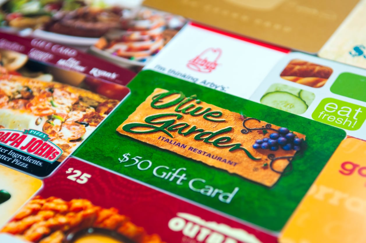 Assorted restaurant gift cards, including Olive Garden, Papa John’s, Arby’s, and Eat Fresh, are shown laid out together. A $50 Olive Garden gift card is in focus at the center.