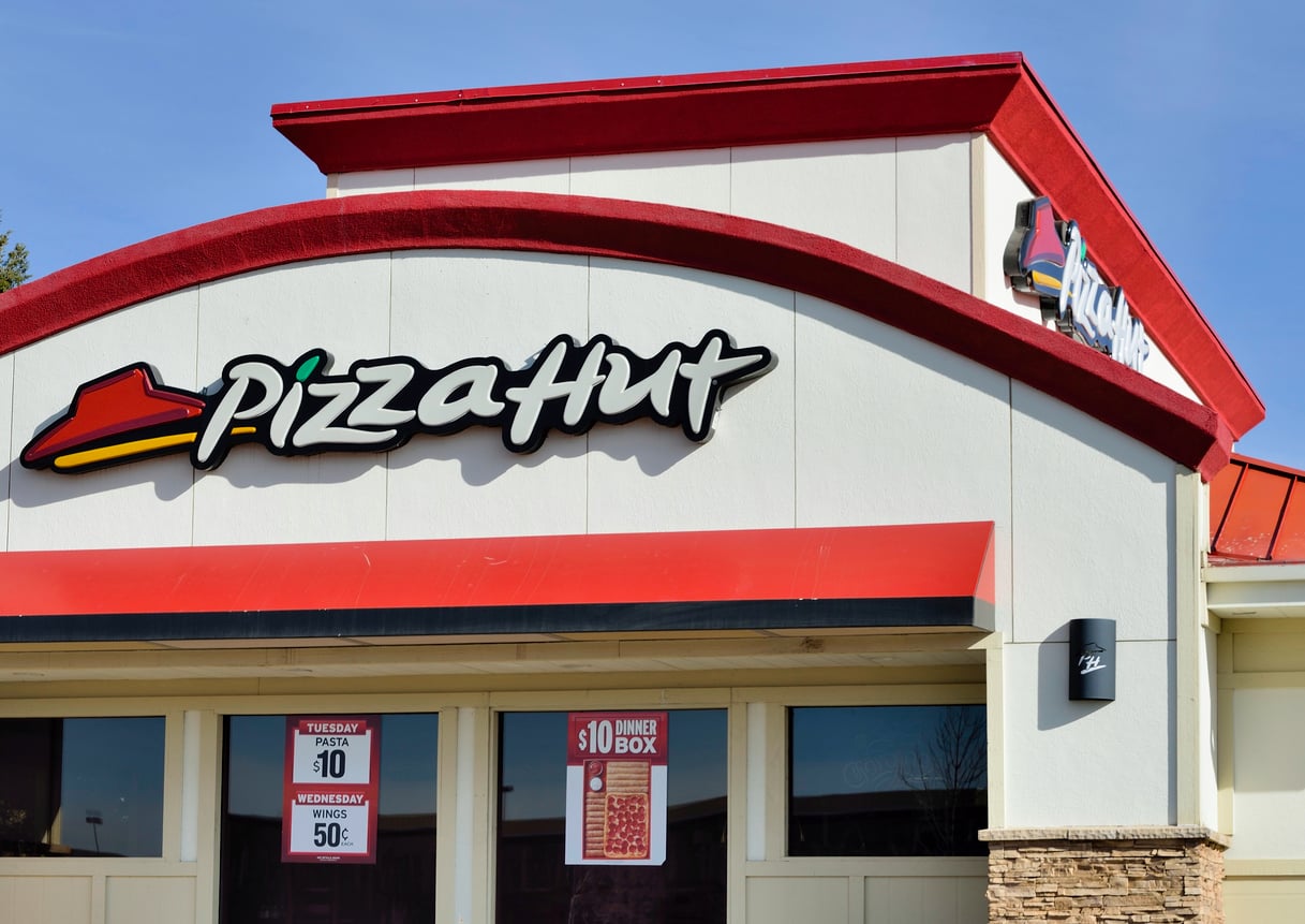 A Pizza Hut restaurant exterior with a red roof, large Pizza Hut logo, and promotional signs advertising deals in the windows.
