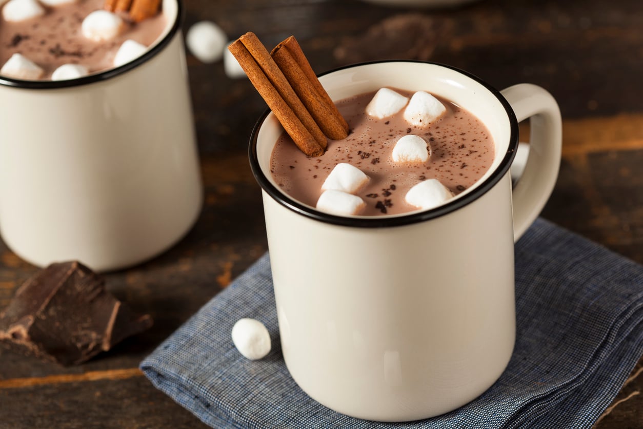 A white mug filled with hot chocolate, topped with mini marshmallows and two cinnamon sticks, sits on a blue napkin with pieces of chocolate nearby on a wooden table.