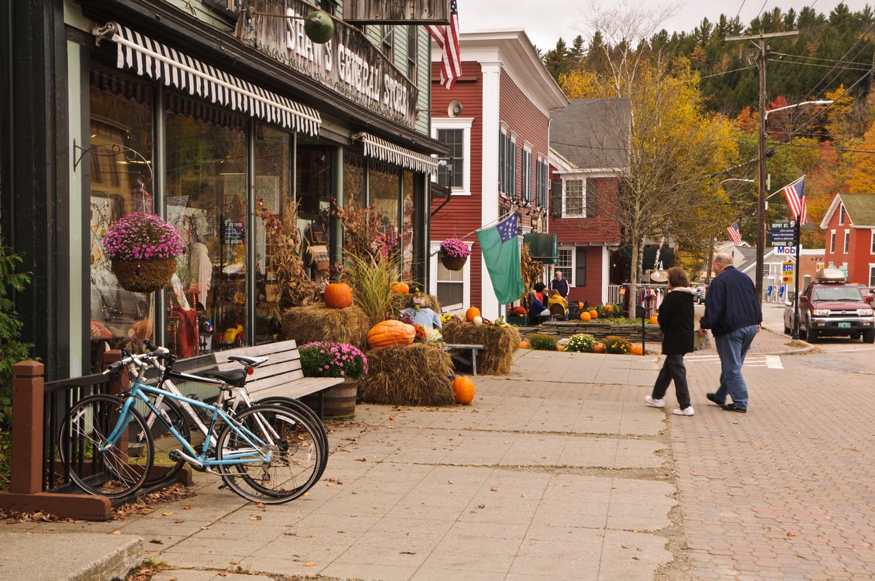 A small town street decorated with pumpkins and hay bales for autumn offers charming vacation experiences. Two people walk on the sidewalk past shops with flowers. Bicycles are parked nearby, and trees with fall foliage line the background.