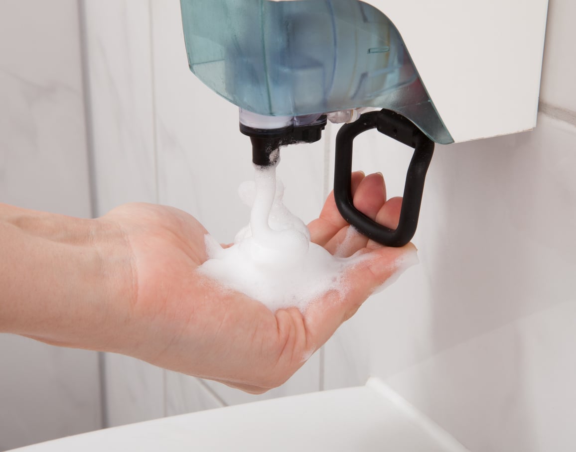 A hand held under a soap dispenser is receiving foamy soap in a bathroom setting with white tiled walls.