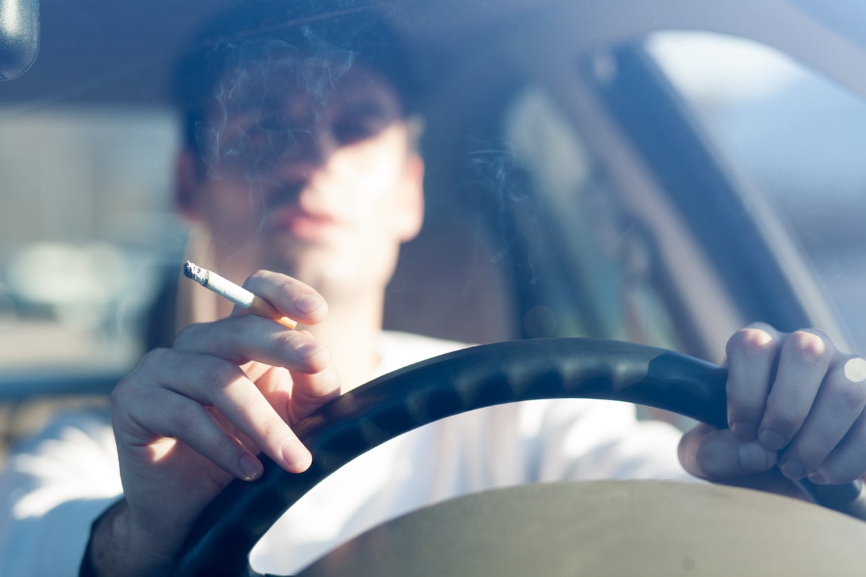 A person sits behind a car steering wheel, perhaps on vacation, holding a lit cigarette with smoke visible. The focus is on the cigarette and hand; the person’s face is blurred and partially obscured by smoke.