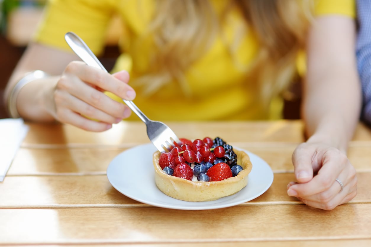 A person in a yellow shirt holds a fork and is about to eat a fruit tart topped with red currants, strawberries, and blueberries on a white plate on a wooden table.