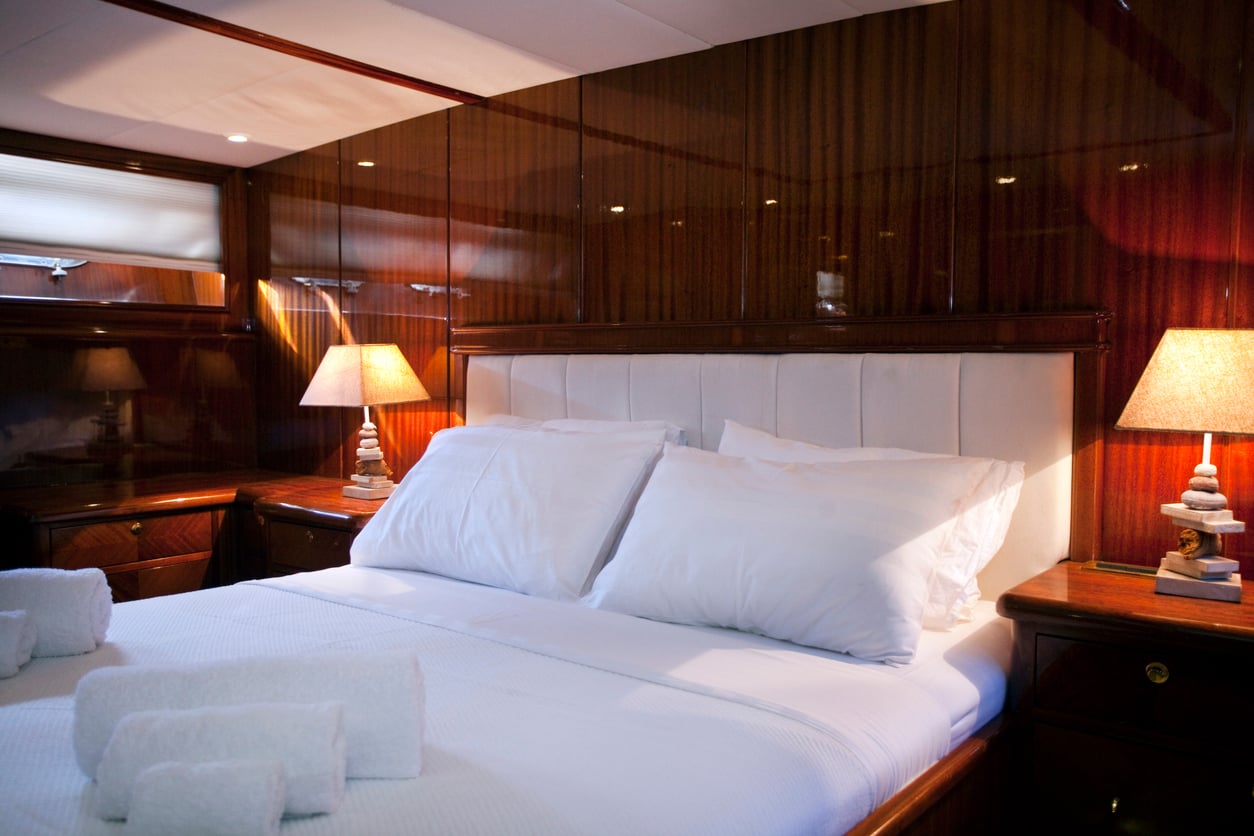 A cozy bedroom with a large bed, white linens, wooden walls, and two bedside tables with lamps. Rolled white towels are placed on the bed, and soft warm lighting creates a relaxing atmosphere.