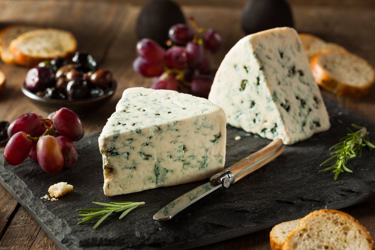 Two wedges of blue cheese on a slate board with a knife, surrounded by grapes, olives, sliced baguette, and sprigs of rosemary on a rustic wooden table—one of those Foods People Learn to Appreciate Later in Life.