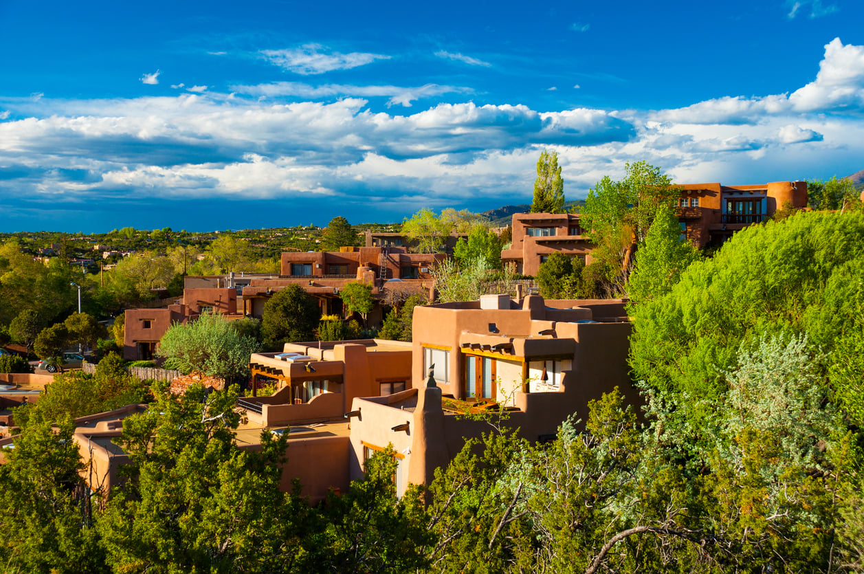 Adobe-style houses surrounded by green trees under a vibrant blue sky with scattered clouds in a Southwestern landscape, likely in Santa Fe, New Mexico.