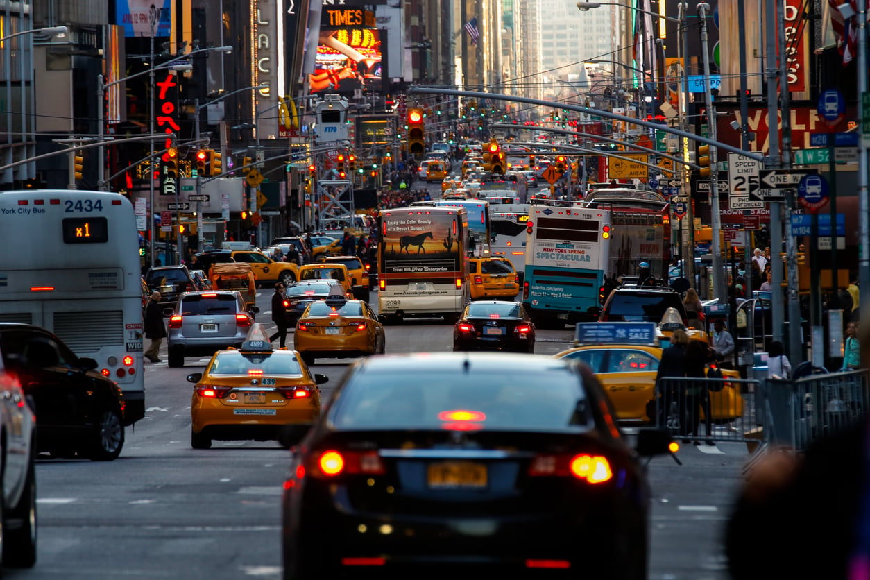 A busy city street packed with yellow taxis, cars, and buses, surrounded by tall buildings and bright neon signs, capturing the bustling atmosphere of an urban downtown area.