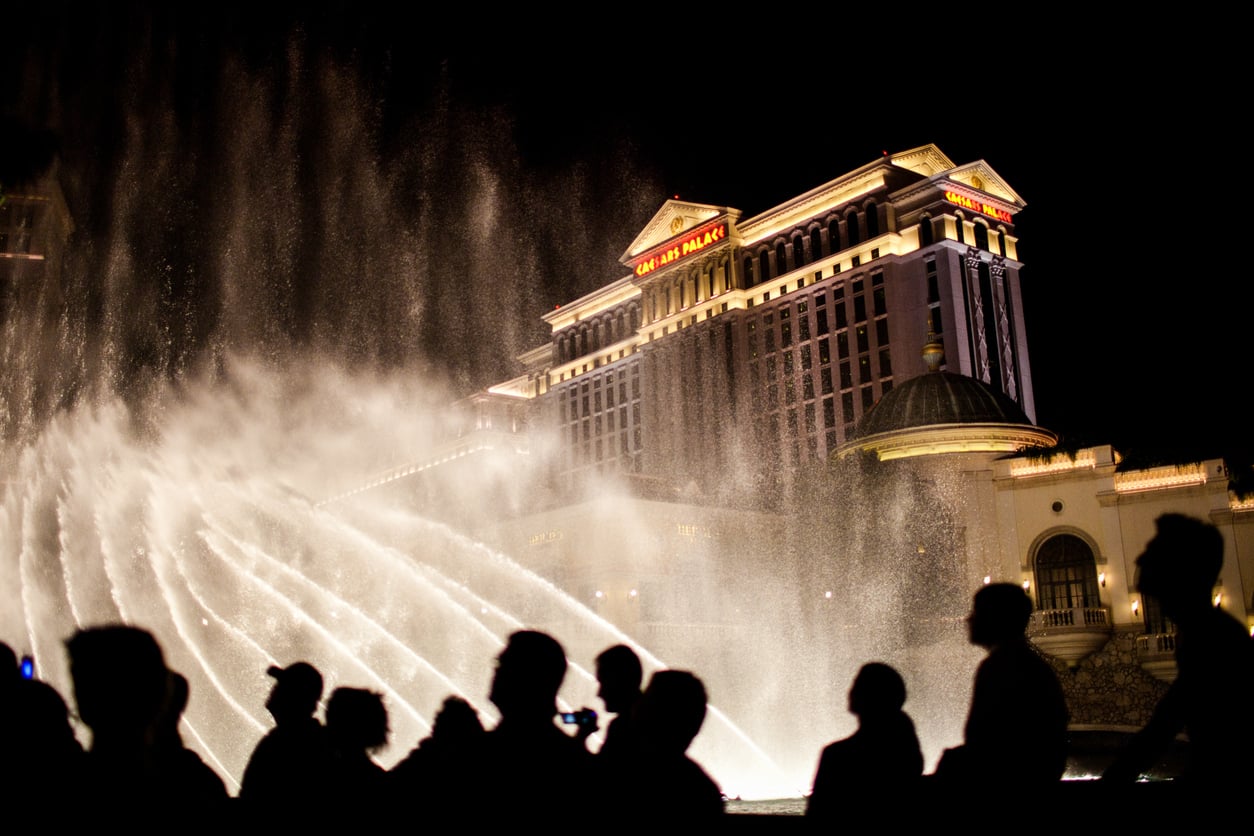 Silhouetted people watch a nighttime fountain show in front of Caesars Palace hotel, brightly lit against the dark sky in Las Vegas.