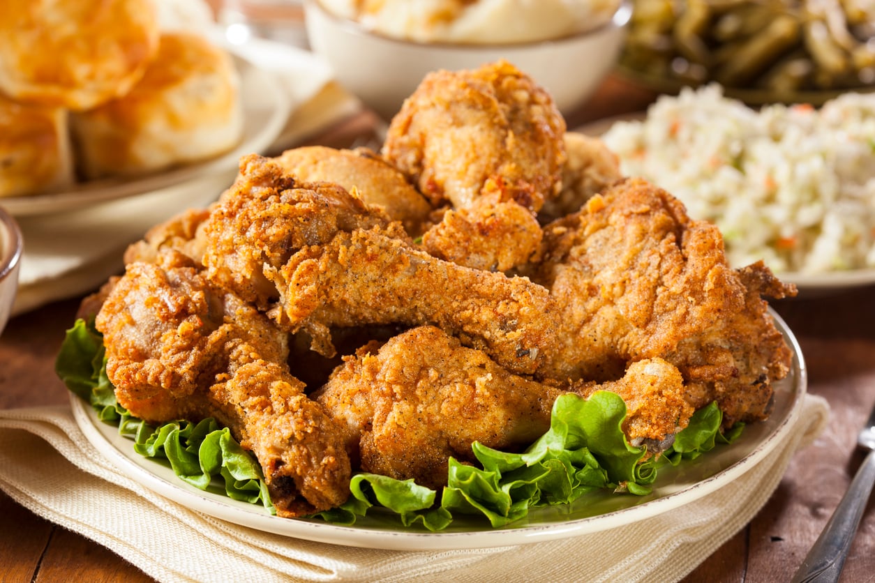 A plate of crispy fried chicken drumsticks sits on a bed of green lettuce, with side dishes like mashed potatoes, biscuits, coleslaw, and green beans visible in the background on a wooden table.