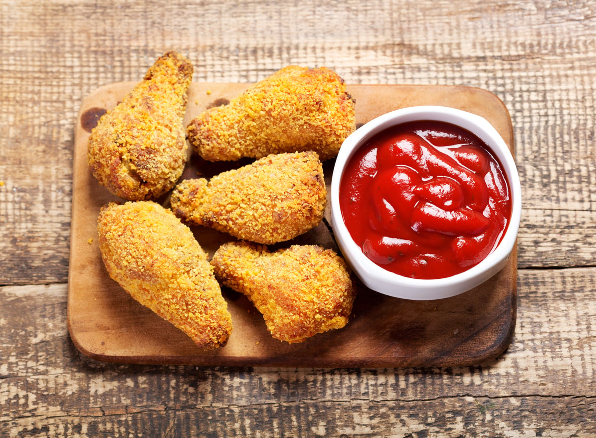 Four crispy fried chicken drumsticks on a wooden board next to a small white bowl filled with ketchup, all set on a rustic wooden table.